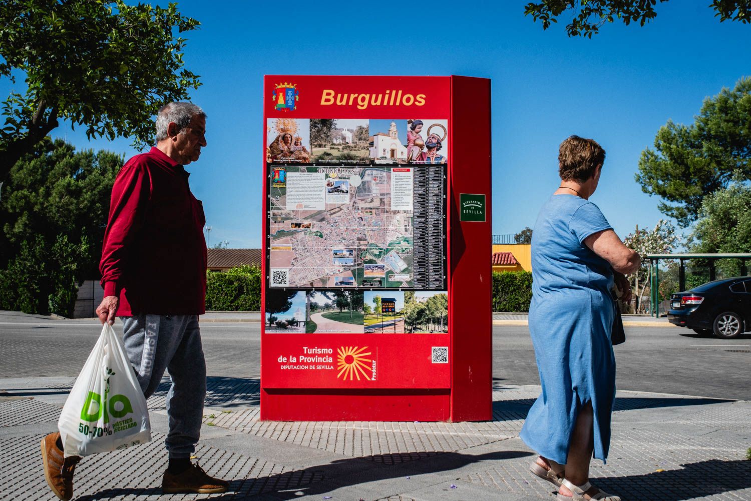 Dos vecinos de Burguillos pasean ante un cartel turístico del municipio.